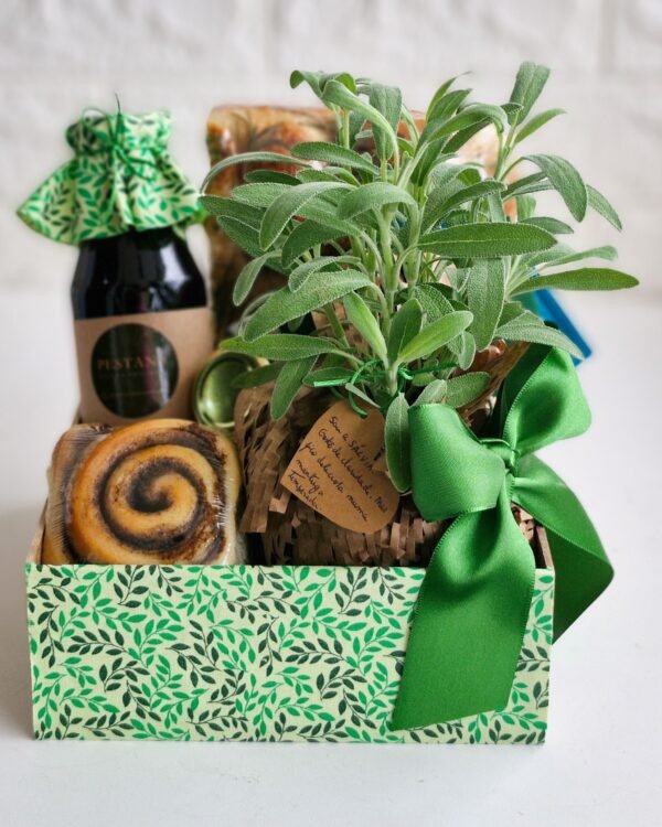 Inicio 85 Cesta de café da manhã Alecrim em caixa rústica com estampa de folhas verdes e elegante laço de cetim verde. Destaque para um vaso de sálvia fresca com tag de papel kraft manuscrita, ao lado de uma focaccia de fermentação longa, rolinho de canela doce e bebida com tampa de tecido. Opção de presente afetivo e botânico em São Gonçalo.