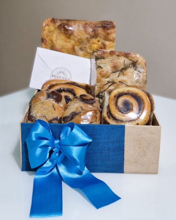 Inicio 84 Cesta de café da manhã em caixa de MDF envolta em tecido azul texturizado com um grande laço de fita de cetim azul bic. Exibe focaccias artesanais de provolone e alecrim, rolinhos de canela, cookies de chocolate e um envelope branco clássico com o selo da Pestana.
