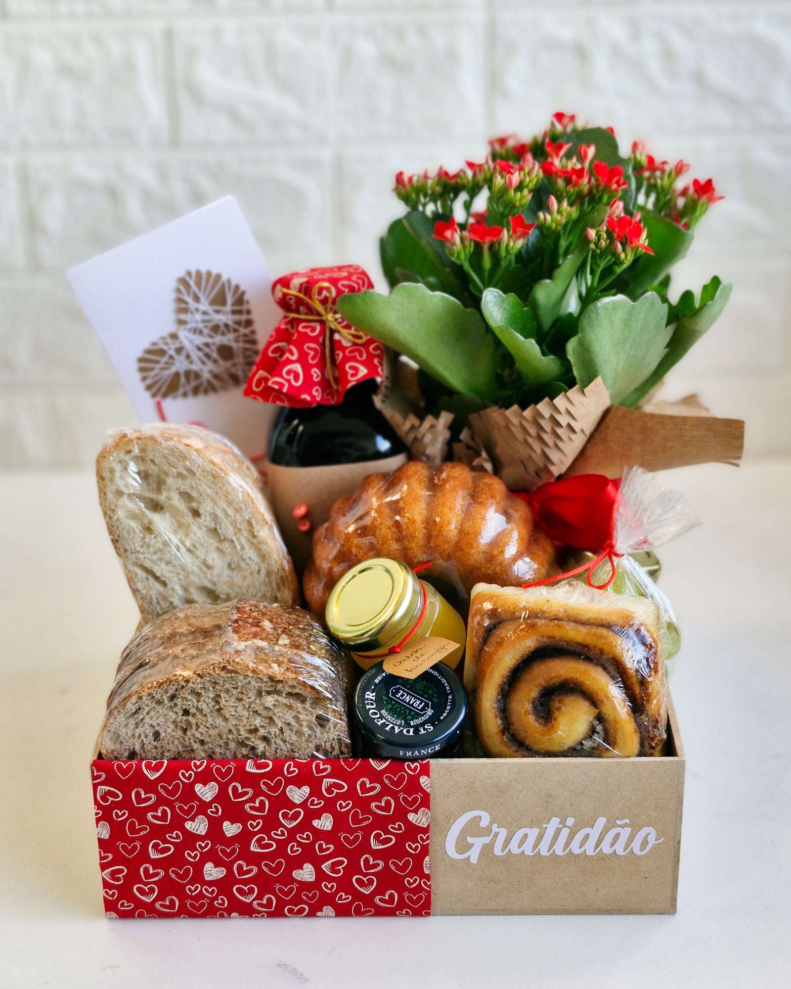 Cesta Encanto 85 Cesta de café da manhã Gratidão em caixa de papel kraft com a palavra 'Gratidão' e faixa estampada com corações vermelhos. A composição inclui fatias de pão rústico de fermentação natural, um minibolo caseiro, rolinho de canela, potes de geleia premium e mel, uma garrafa artesanal com tampa de tecido vermelho, um cartão afetivo e um vaso de flores vermelhas naturais (Kalanchoe). Opção de presente afetivo em São Gonçalo.