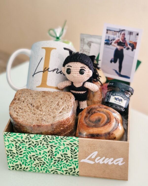 Inicio 82 Cesta de café da manhã fitness premium em caixa de MDF com estampa de folhas verdes e o nome 'Luna'. A composição exclusiva exibe uma boneca amigurumi em crochê com roupas de ginástica pretas, foto polaroid de uma mulher correndo, caneca personalizada, fatias de pão 100% integral, rolinho de canela e geleia de mirtilo sem açúcar.