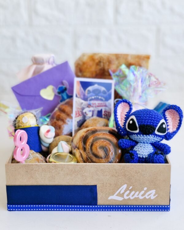 Inicio 80 Cesta de café da manhã infantil em caixa de MDF decorada com fita azul e o nome 'Lívia' em branco. Em destaque, um boneco amigurumi em crochê do personagem Stitch, uma foto revelada da criança, um envelope lilás temático, vela rosa de aniversário número 8, bombom Ferrero Rocher, marshmallows coloridos, rolinho de canela e pães artesanais rústicos