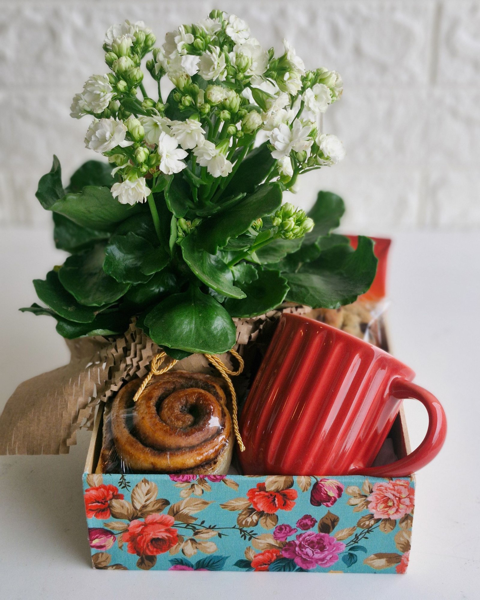 Quem Somos 2 Cesta de café da manhã em caixa de MDF com estampa floral em tons de azul, vermelho e rosa. Contém uma caneca de cerâmica vermelha texturizada, flor Kalanchoe branca em embalagem rústica de papel kraft, rolinho de canela e deliciosos biscoitos artesanais ao fundo.