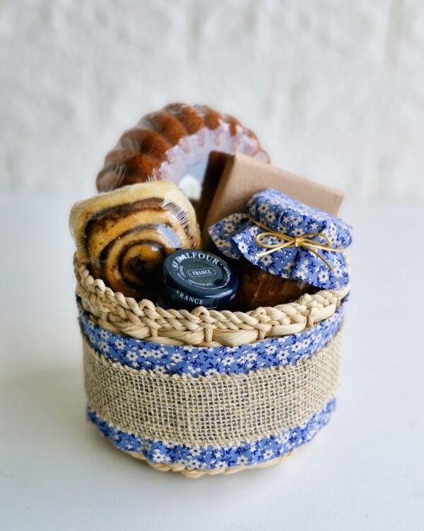 Inicio 79 Cesta de café da manhã em cesto de taboa decorado com tecido floral azul e faixa de juta rústica. A composição farta contém um mini bolo de laranja caseiro, rolinho de canela macio, geleia francesa St. Dalfour, caixa kraft e um potinho de vidro com tampa de tecido floral azul combinando com a embalagem.