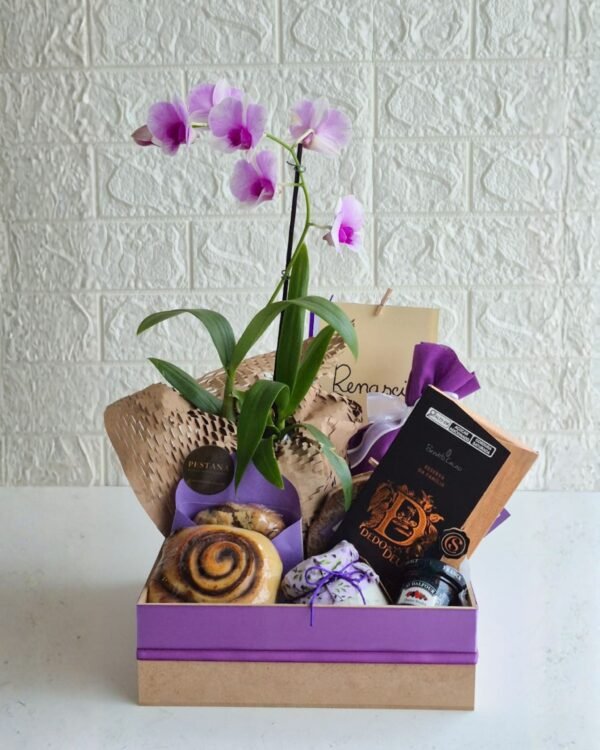 Inicio 78 Cesta de café da manhã premium em caixa de MDF com base crua e tampa roxa. Em destaque, uma orquídea lilás alta envolvida em papel kraft sustentável favo de mel. A composição inclui barra de chocolate Dedo de Deus, rolinho de canela, cookie artesanal em envelope roxo com selo Pestana, potinho de pasta salgada com tampa de tecido floral roxo, geleia francesa St. Dalfour e cartão.