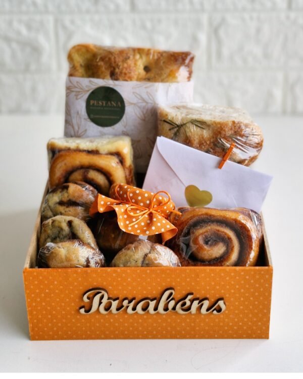 Inicio 81 Cesta de café da manhã de aniversário em caixa laranja com a palavra "Parabéns" em madeira. Contém pães artesanais, biscoitos, geleia e um cartão com coração.