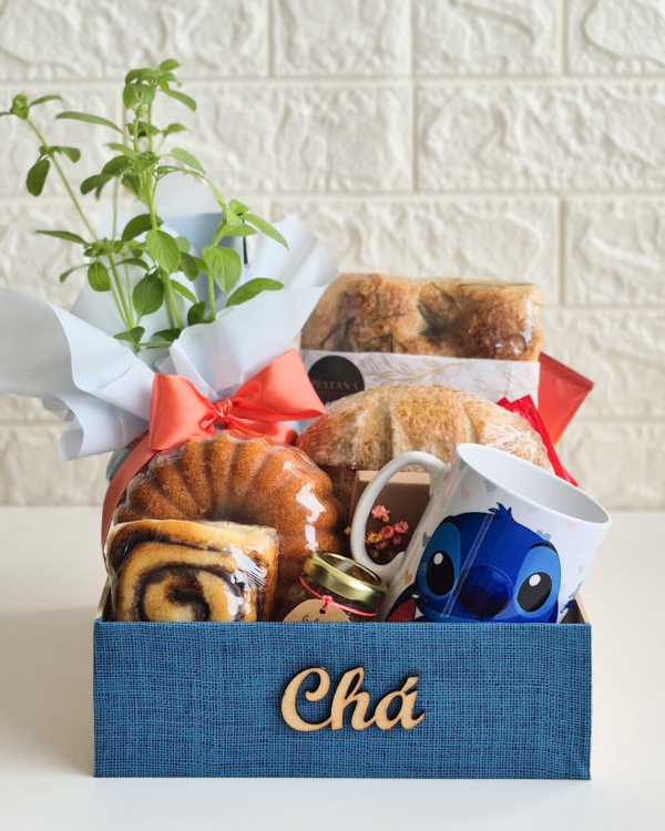 Nossas Cestas 3 Cesta de chá da tarde ou café da manhã em caixa azul texturizada com a palavra "Chá" em madeira. A cesta contém uma caneca personalizada do personagem Stitch (Disney), um vaso de manjericão fresco embalado para presente com laço laranja, pães rústicos de fermentação natural, um bolo caseiro redondo, rolinho doce e um pote de vidro pequeno (geleia/mel).