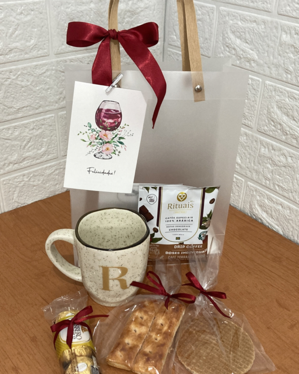 Bag Afeto transparente com alça rústica, laço vermelho e cartão Felicidades. À frente, xícara com inicial R, café drip coffee, biscoitos finos e chocolates Ferrero Rocher sobre mesa de madeira.