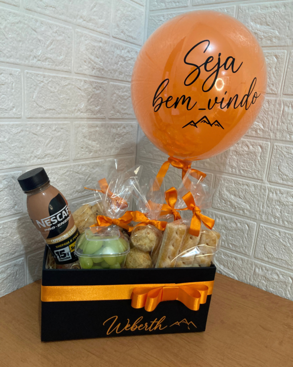 Box Legendário em caixa preta com laço laranja, personalizada com o nome Weberth. Contém Nescafé, uvas frescas, croissant, pães de queijo e biscoitos, além de um balão laranja escrito Seja bem-vindo.