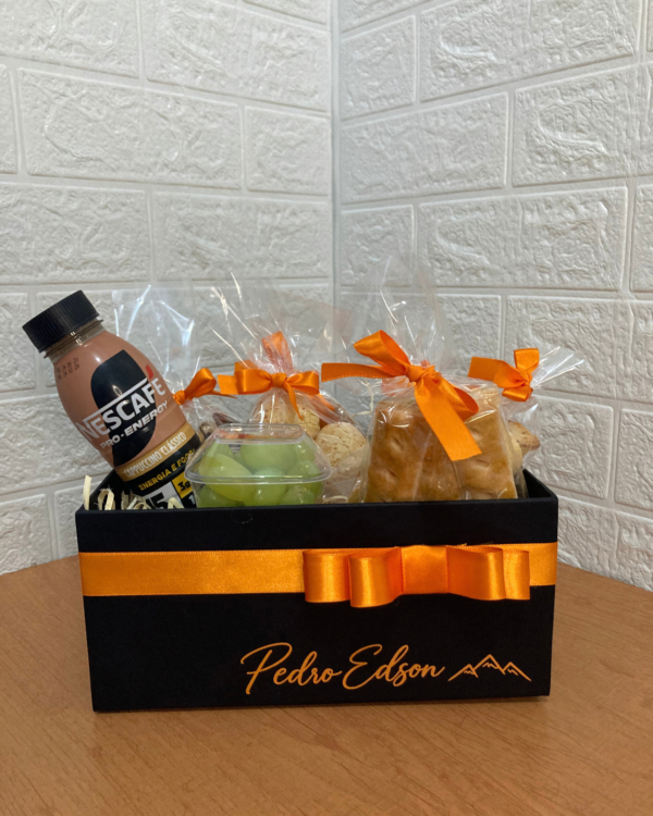 Cesta Box Legendário 5 Box Legendário em caixa preta com o nome Pedro Edson, fita laranja, contendo Nescafé, uvas e embalagens de biscoitos e pães de queijo.