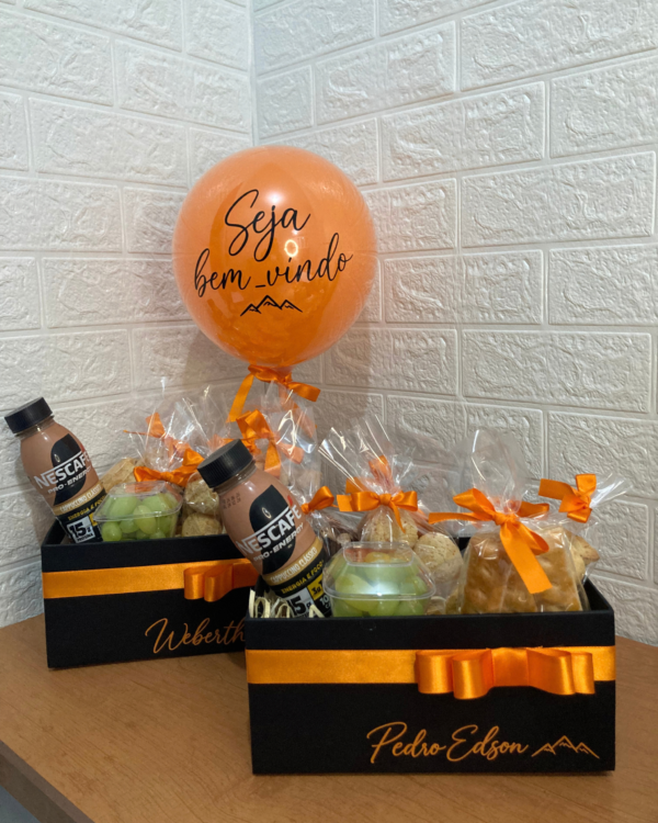 Cesta Box Legendário 2 Duas unidades da Box Legendário em caixas pretas com laços laranjas, personalizadas com os nomes Weberth e Pedro Edson, recheadas com café, uvas e pães.