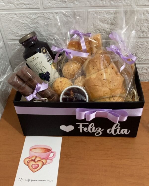 Cesta de café da manhã em caixa preta decorada com a frase Feliz dia e fita lilás. O interior contém uma garrafa de suco de uva Casa Madeira, pães de queijo, pães rústicos e biscoitos de chocolate, todos embalados individualmente com laços. À frente da cesta, há um cartão com ilustração de uma xícara rosa e a frase "Um café para comemorar!