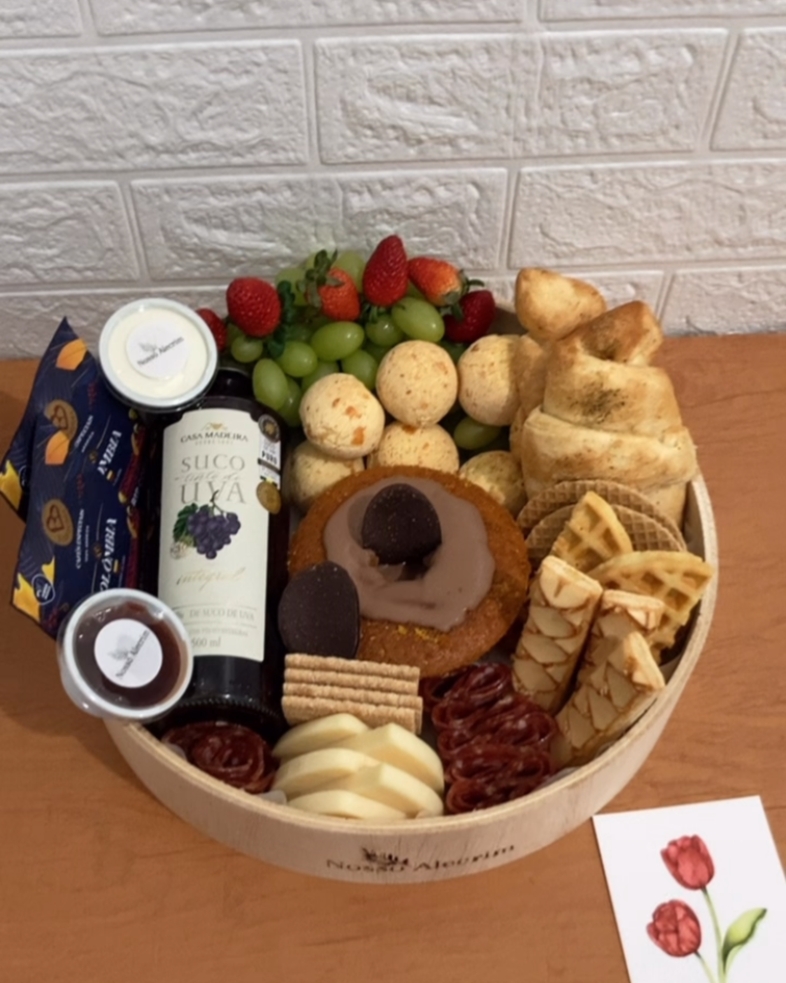 Visão superior da Cesta G na bandeja de madeira com suco de uva, café, frutas frescas, pães, frios, biscoitos e bolo de cenoura com chocolate.