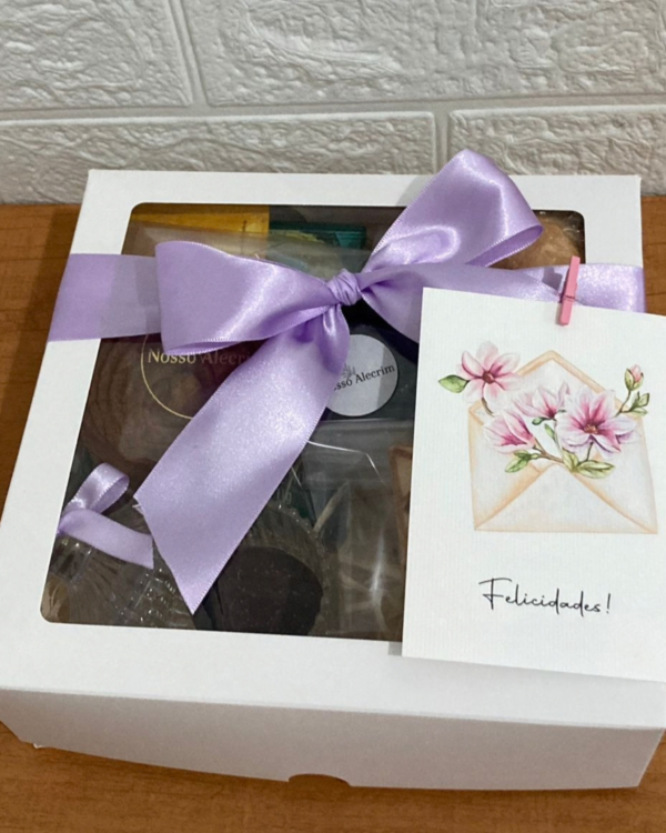 Cesta Café Mimo 4 Cesta de café da manhã em caixa branca com laço lilás e cartão com ilustração de flores rosas e a mensagem Felicidades, presa por um mini prendedor.
