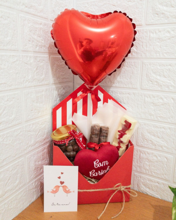 Cesta Chocolate 3 Cesta de chocolates em caixa vermelha formato envelope com um balão metalizado de coração vermelho. Contém um pote de vidro com bombons, barras de chocolate, uma pelúcia de coração com a frase Com Carinho e um cartão com pássaros e a mensagem Eu te amo!