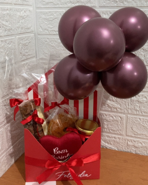 Cesta Chocolate 2 Cesta de chocolate da Nosso Alecrim em caixa envelope vermelha com a frase "Feliz dia" e laço de cetim vermelho. No interior, guloseimas embaladas, um pote com tampa dourada, coração de pelúcia com a frase "Com Carinho", cartão "Parabéns!" com desenho de bolo e um arranjo de balões metalizados na cor vinho, apoiada em mesa de madeira.