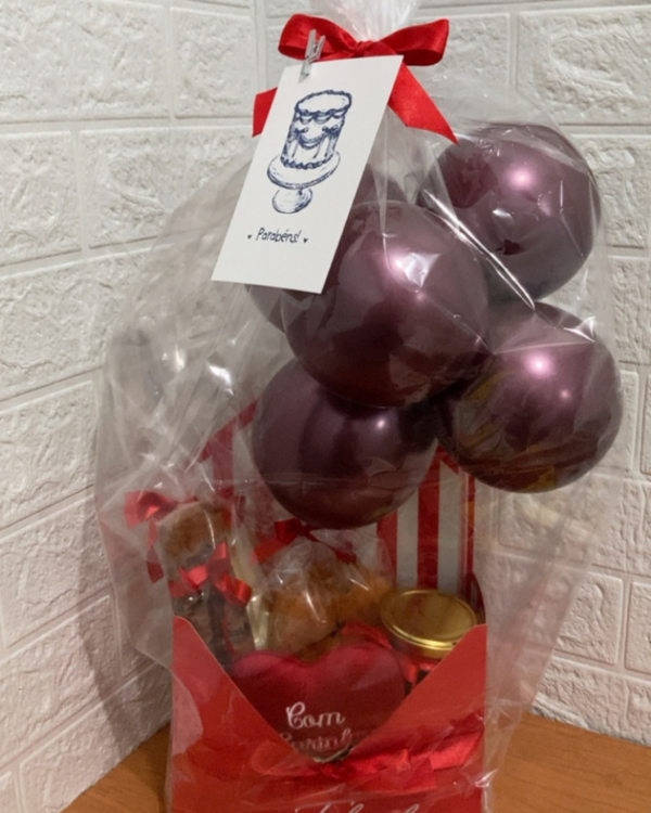 Cesta Chocolate 4 Cesta de chocolate da Nosso Alecrim embalada para presente em celofane transparente, fechada com laço de cetim vermelho e um cartão "Parabéns!" preso com prendedor no topo. Visível em seu interior a caixa envelope vermelha "Feliz dia", coração de pelúcia, pote, guloseimas e balões metalizados na cor vinho, sobre mesa de madeira.