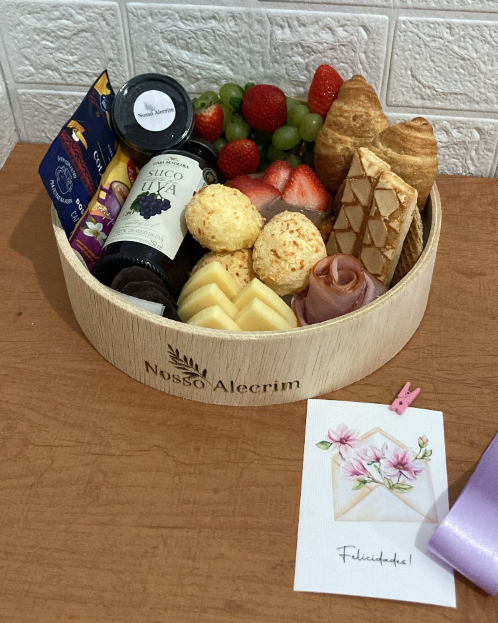 Cesta G 3 Cesta G em bandeja de madeira repleta de pães, frios, frutas e suco, acompanhada em primeiro plano por um cartão com desenho de envelope floral escrito Felicidades.