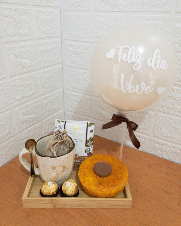 Bandeja de bambu com uma caneca rústica de cerâmica, um sachê de drip coffee artesanal, um minibolo caseiro com uma moeda de chocolate no topo e dois bombons Ferrero Rocher. Ao fundo, um balão branco personalizado com a frase Feliz dia Vovó e um laço castanho