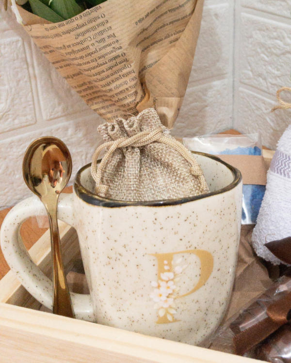 Cesta Ritual de Carinho 2 Detalhe da xícara de porcelana rústica com a inicial P em dourado, acompanhada por uma colher dourada e um pequeno saquinho de juta no interior.