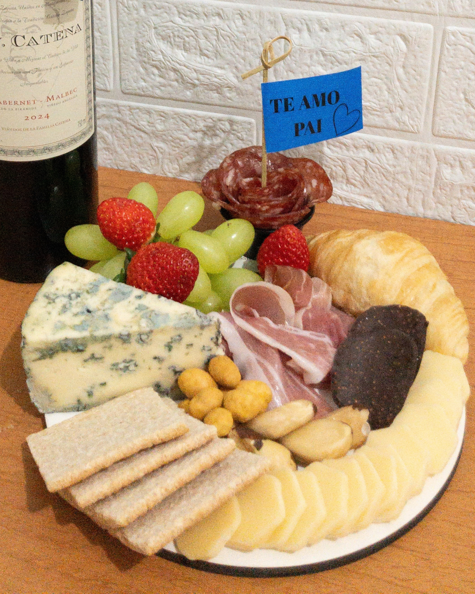 Tábua de Frios Momento 3 Plano aproximado da Tábua Momento destacando o queijo azul, presunto de Parma, uvas, morangos, flor de salame com plaquinha Te Amo Pai e parte da garrafa de vinho D.V. Catena.
