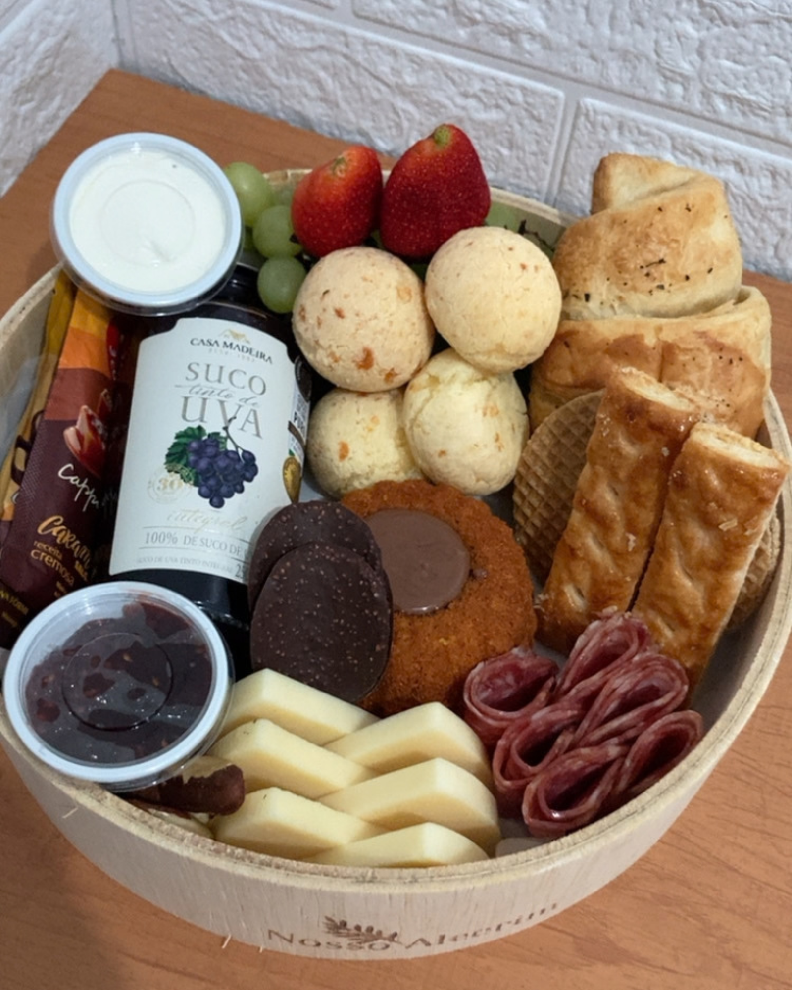 Cesta P 4 Vista aproximada dos ingredientes da Cesta P, destacando o bolo de cenoura, sachê de cappuccino, suco, pães de queijo, frios e frutas vermelhas
