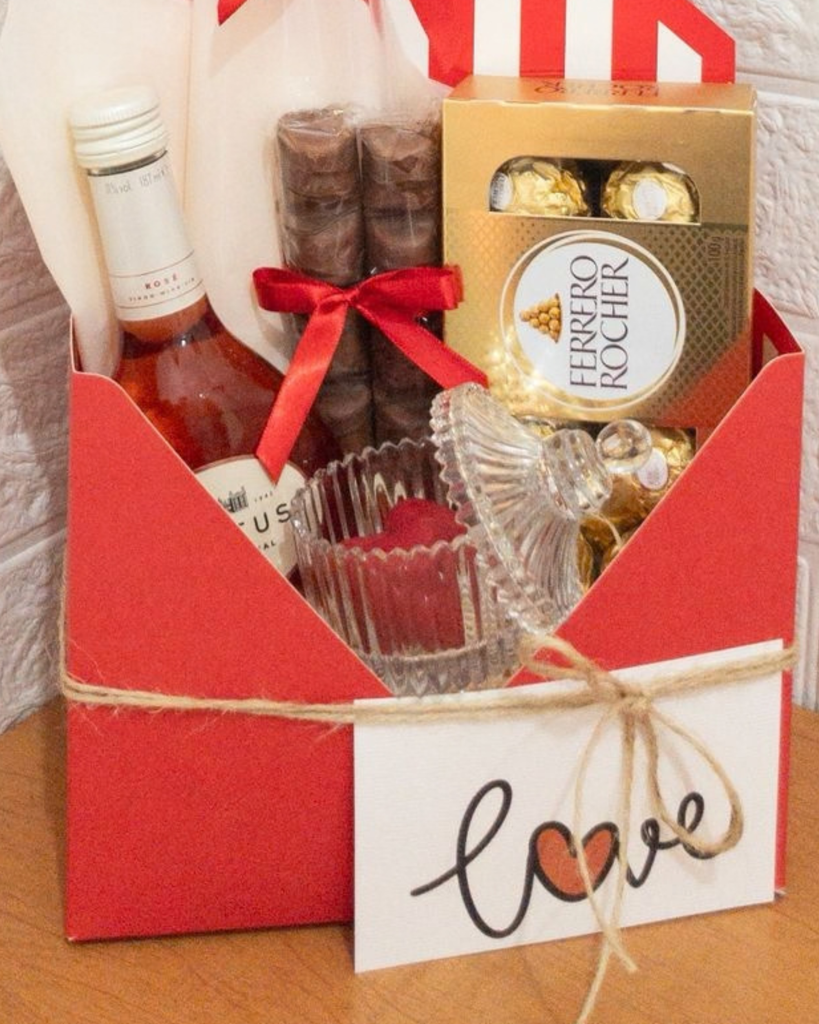 Cesta de Vinho e Chocolate 3 Detalhe da caixa envelope vermelha com vinho rosé, chocolates Kinder Bueno, caixa de Ferrero Rocher e bomboniere de vidro aberta com chocolate de coração.
