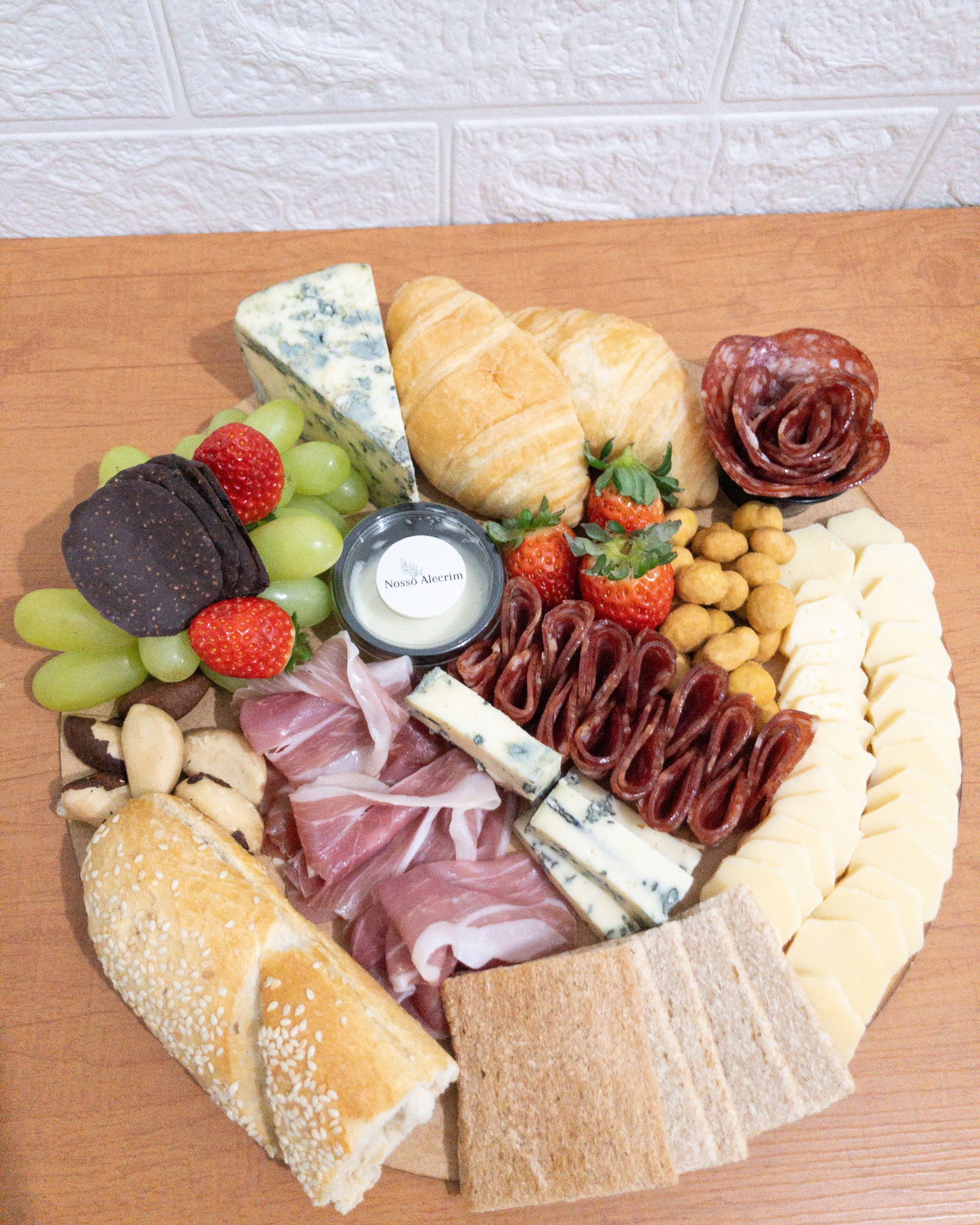 Tábua de Frios Momento 4 Visão superior de uma tábua de frios muito farta com baguete, croissant, queijo azul, fatias de queijo, presunto de Parma, salame, uvas, morangos, castanhas e potinho da Nosso Alecrim.